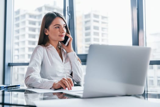 Have conversation by the phone. Young woman in white formal clothes is indoors in the modern office.