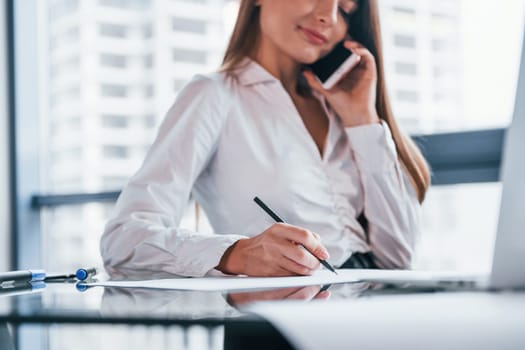Have conversation by the phone. Young woman in white formal clothes is indoors in the modern office.