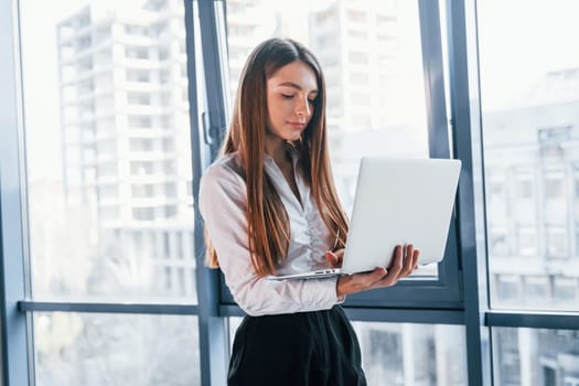 Standing with laptop. Young woman in white formal clothes is indoors in the modern office.