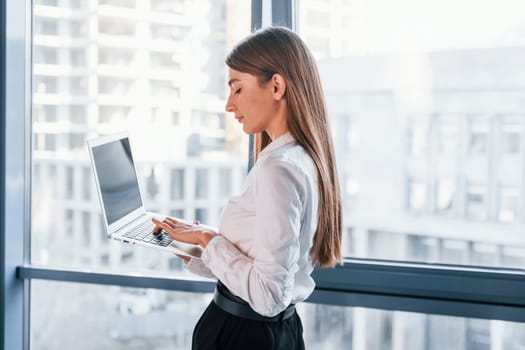 Standing with laptop. Young woman in white formal clothes is indoors in the modern office.