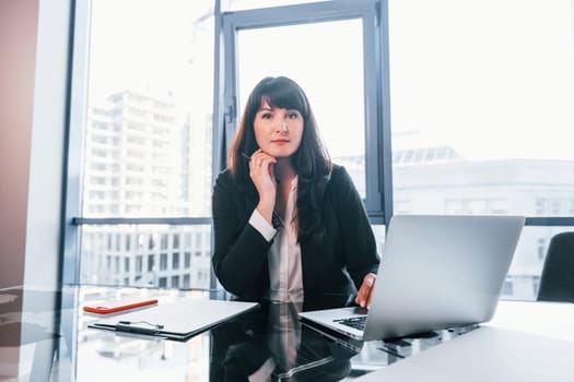 With laptop on table. Woman in black formal clothes is indoors in the modern office.