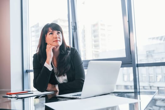 With laptop on table. Woman in black formal clothes is indoors in the modern office.