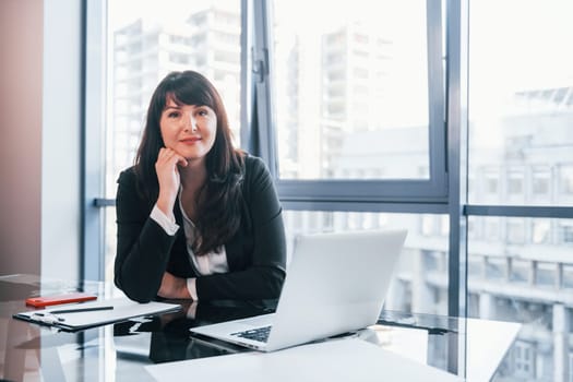 With laptop on table. Woman in black formal clothes is indoors in the modern office.