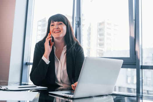 With laptop on table. Woman in black formal clothes is indoors in the modern office.