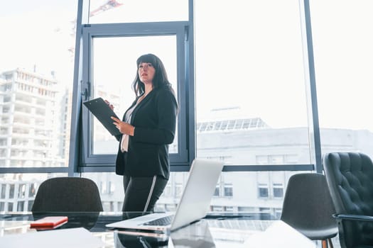 With laptop on table. Woman in black formal clothes is indoors in the modern office.