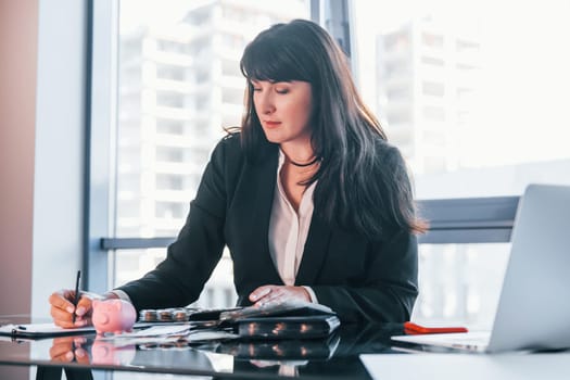With laptop on table. Woman in black formal clothes is indoors in the modern office.
