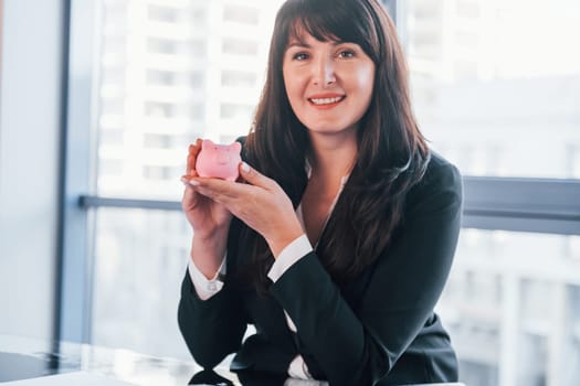 Woman in black formal clothes is indoors in the modern office holds pink piggy bank.