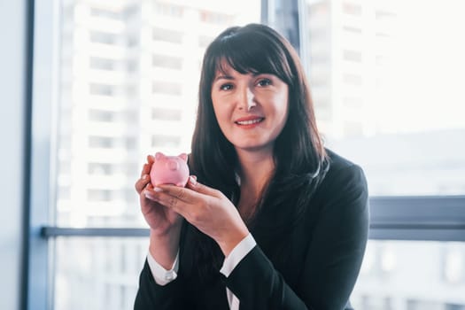 Woman in black formal clothes is indoors in the modern office holds pink piggy bank.