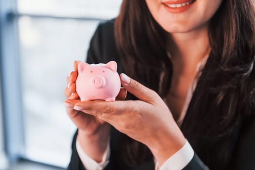 Woman in black formal clothes is indoors in the modern office holds pink piggy bank.