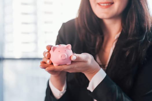 Woman in black formal clothes is indoors in the modern office holds pink piggy bank.