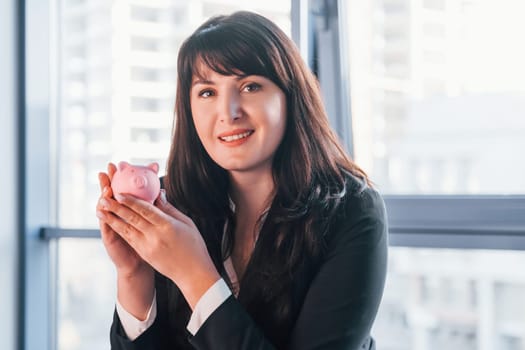 Woman in black formal clothes is indoors in the modern office holds pink piggy bank.