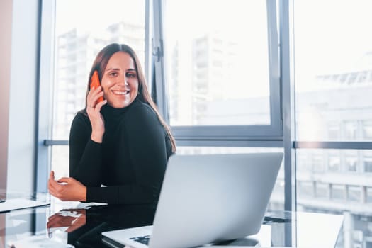 With laptop on table. Woman in black formal clothes is indoors in the modern office.