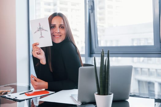 With laptop on table. Woman in black formal clothes is indoors in the modern office.