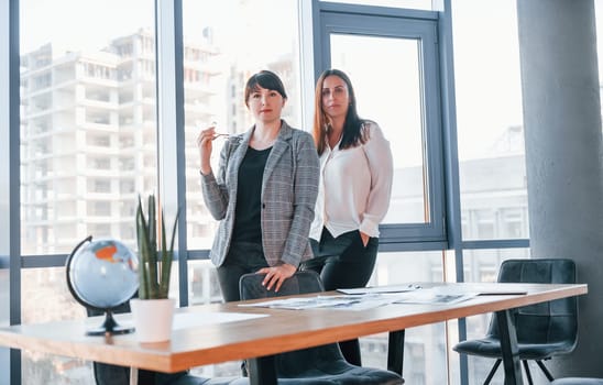 Two women in formal clothes is indoors in the modern office works together.