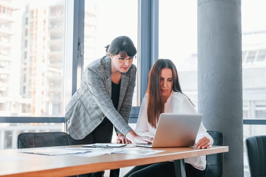 Using laptop. Two women in formal clothes is indoors in the modern office works together.