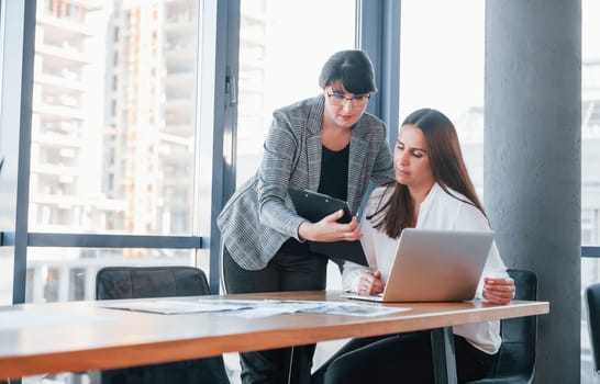 Using laptop. Two women in formal clothes is indoors in the modern office works together.