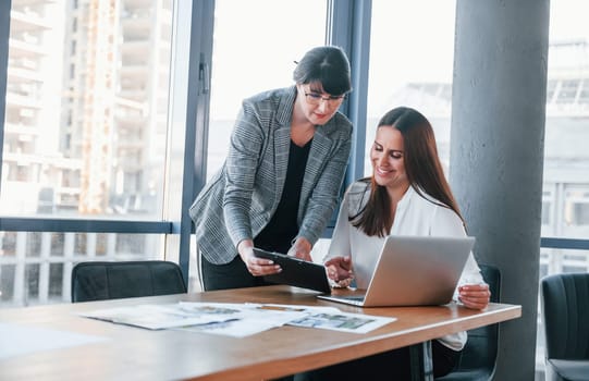 Using laptop. Two women in formal clothes is indoors in the modern office works together.