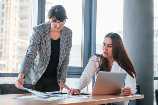 Using laptop. Two women in formal clothes is indoors in the modern office works together.
