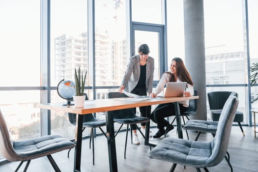 Using laptop. Two women in formal clothes is indoors in the modern office works together.
