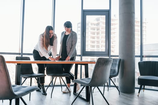 Standing and talking. Two women in formal clothes is indoors in the modern office works together.
