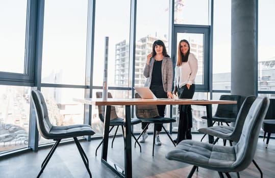 Standing and talking. Two women in formal clothes is indoors in the modern office works together.