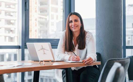 Sits by the table. Young woman in white formal clothes is indoors in the modern office.