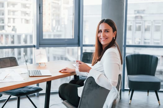 Sits by the table. Young woman in white formal clothes is indoors in the modern office.