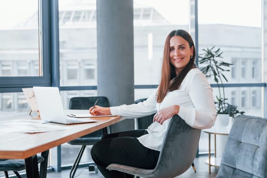 Sits by the table. Young woman in white formal clothes is indoors in the modern office.