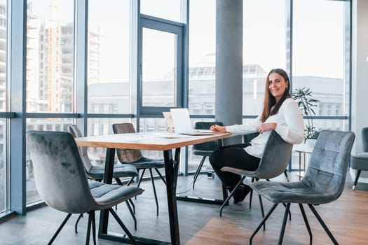 Sits by the table. Young woman in white formal clothes is indoors in the modern office.