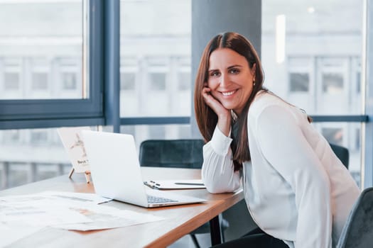 Sits by the table. Young woman in white formal clothes is indoors in the modern office.