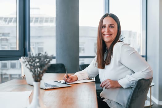 Sits by the table. Young woman in white formal clothes is indoors in the modern office.