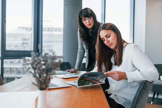 Two women in formal clothes is indoors in the modern office works together.