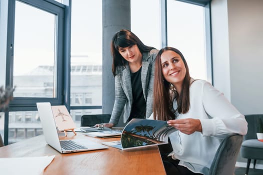 Two women in formal clothes is indoors in the modern office works together.