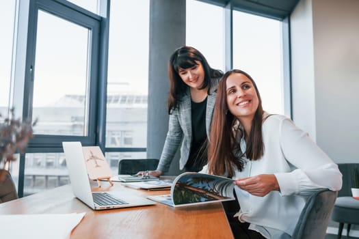 Two women in formal clothes is indoors in the modern office works together.