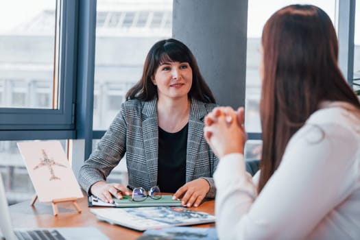 Sitting by the table. Two women in formal clothes is indoors in the modern office works together.