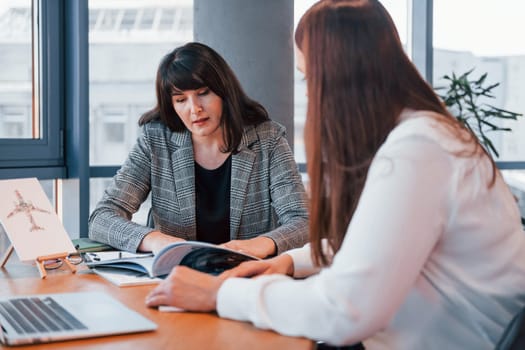 Sitting by the table. Two women in formal clothes is indoors in the modern office works together.