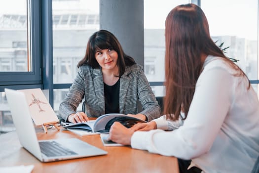 Sitting by the table. Two women in formal clothes is indoors in the modern office works together.