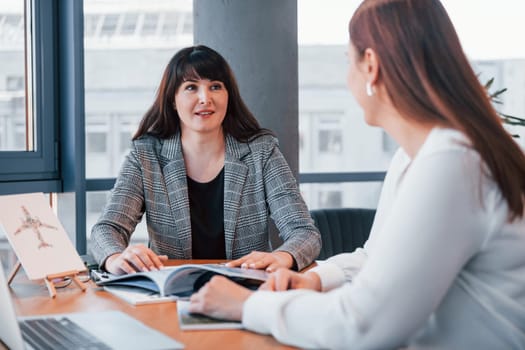 Sitting by the table. Two women in formal clothes is indoors in the modern office works together.