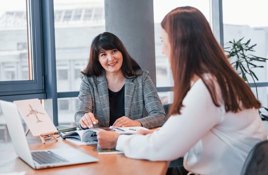 Sitting by the table. Two women in formal clothes is indoors in the modern office works together.