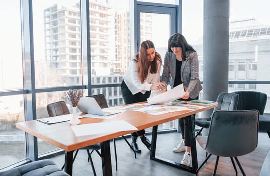 Standing and talking. Two women in formal clothes is indoors in the modern office works together.