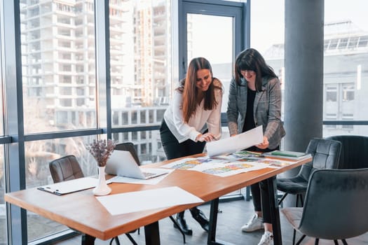 Standing and talking. Two women in formal clothes is indoors in the modern office works together.