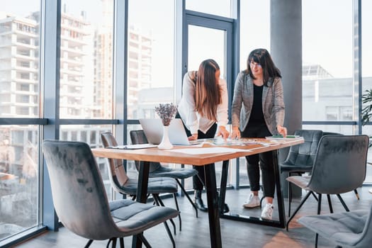 Standing and talking. Two women in formal clothes is indoors in the modern office works together.