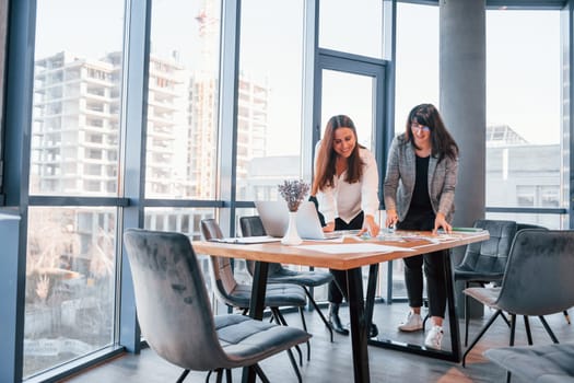 Standing and talking. Two women in formal clothes is indoors in the modern office works together.