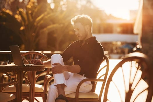 Sits by the table. Young man is outdoors at sunny daytime. Concept of vacation.