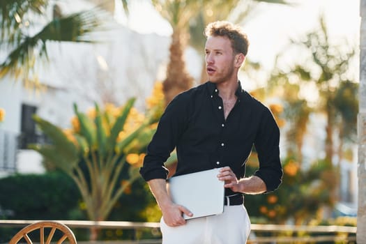 Holds laptop. Young man is outdoors at sunny daytime. Concept of vacation.