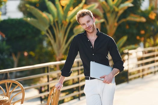 Holds laptop. Young man is outdoors at sunny daytime. Concept of vacation.