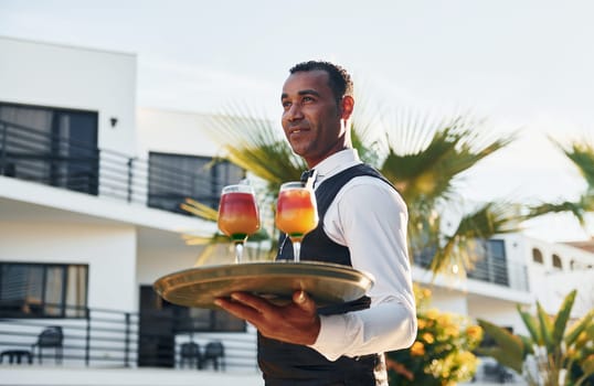 Holds cocktails. Black waiter in formal clothes is at his work outdoors at sunny daytime.