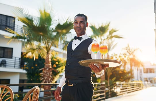 Holds cocktails. Black waiter in formal clothes is at his work outdoors at sunny daytime.