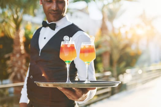 Holds cocktails. Black waiter in formal clothes is at his work outdoors at sunny daytime.