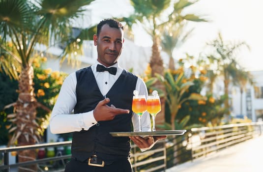 Holds cocktails. Black waiter in formal clothes is at his work outdoors at sunny daytime.